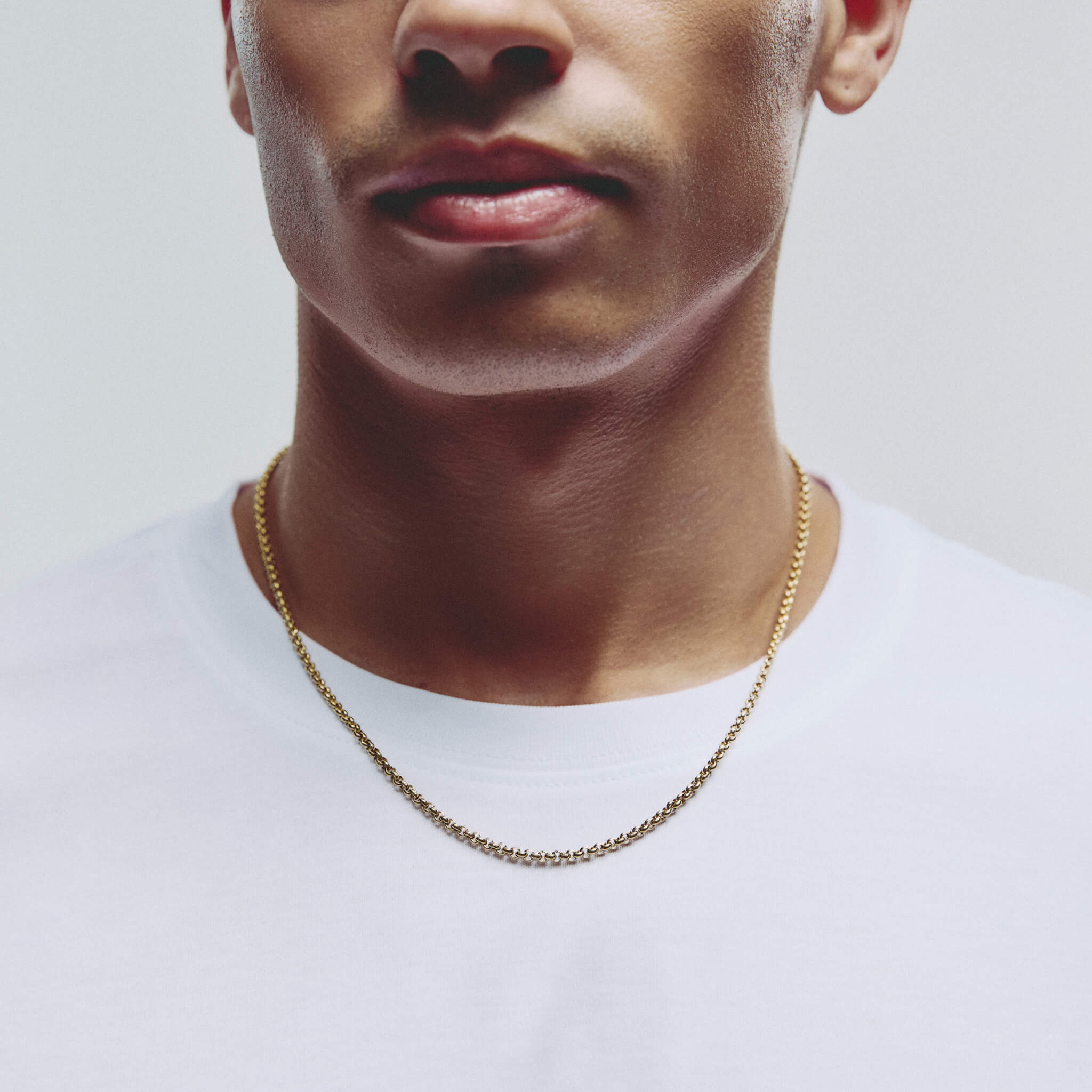 A close-up of a man wearing a delicate gold chain necklace over a white t-shirt, cropped from the nose down against a light gray background.