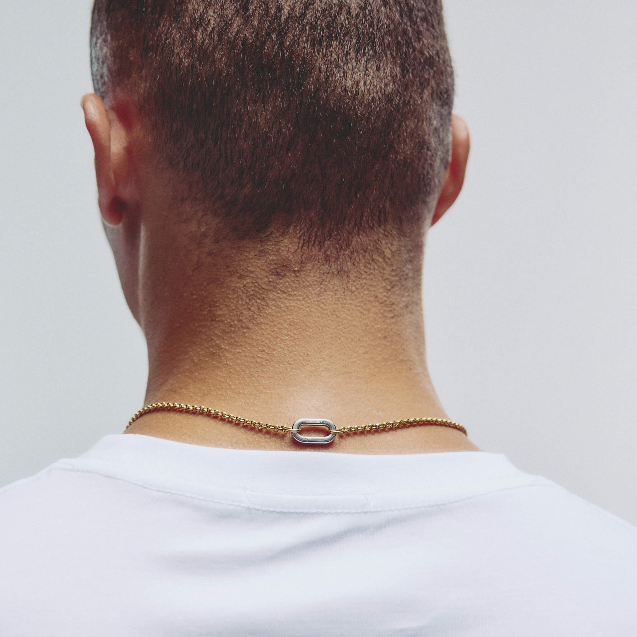 A close-up back view of a person wearing a delicate gold chain necklace with a silver-toned carabiner clasp, paired with a white t-shirt against a light gray background.
