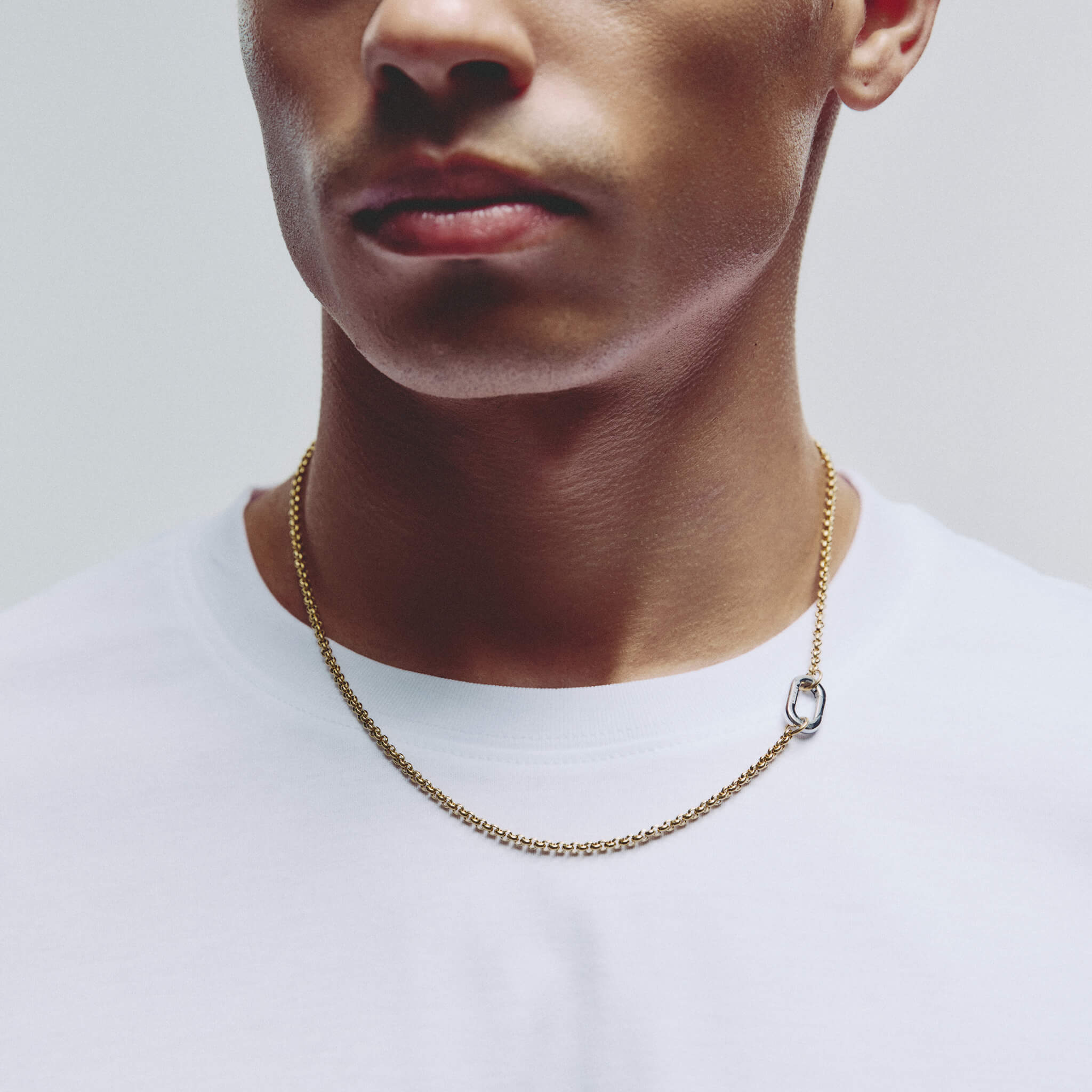 A close-up of a man wearing a delicate gold chain necklace with a small silver carabiner clasp, paired with a white crew-neck t-shirt against a light gray background.