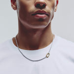 A close-up of a man wearing a silver chain necklace with a small gold carabiner clasp styled with a white crew-neck t-shirt against a light gray background.