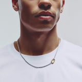 A close-up of a man wearing a silver chain necklace with a small gold carabiner clasp styled with a white crew-neck t-shirt against a light gray background.