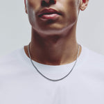 A close-up of a man's neck and lower face wearing a silver chain necklace over a white t-shirt, set against a plain light gray background.