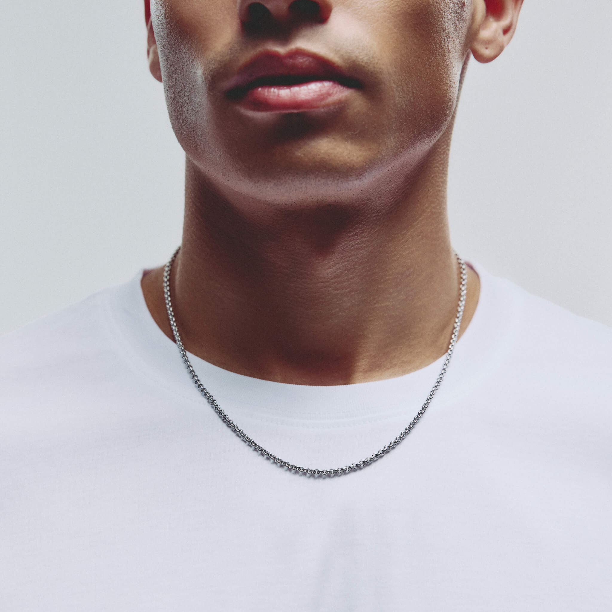 A close-up of a man's neck and lower face wearing a silver chain necklace over a white t-shirt, set against a plain light gray background.