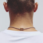 A close-up back view of a man's neck and upper shoulders, wearing a silver-toned necklace with a gold carabiner clasp, against a light gray background.