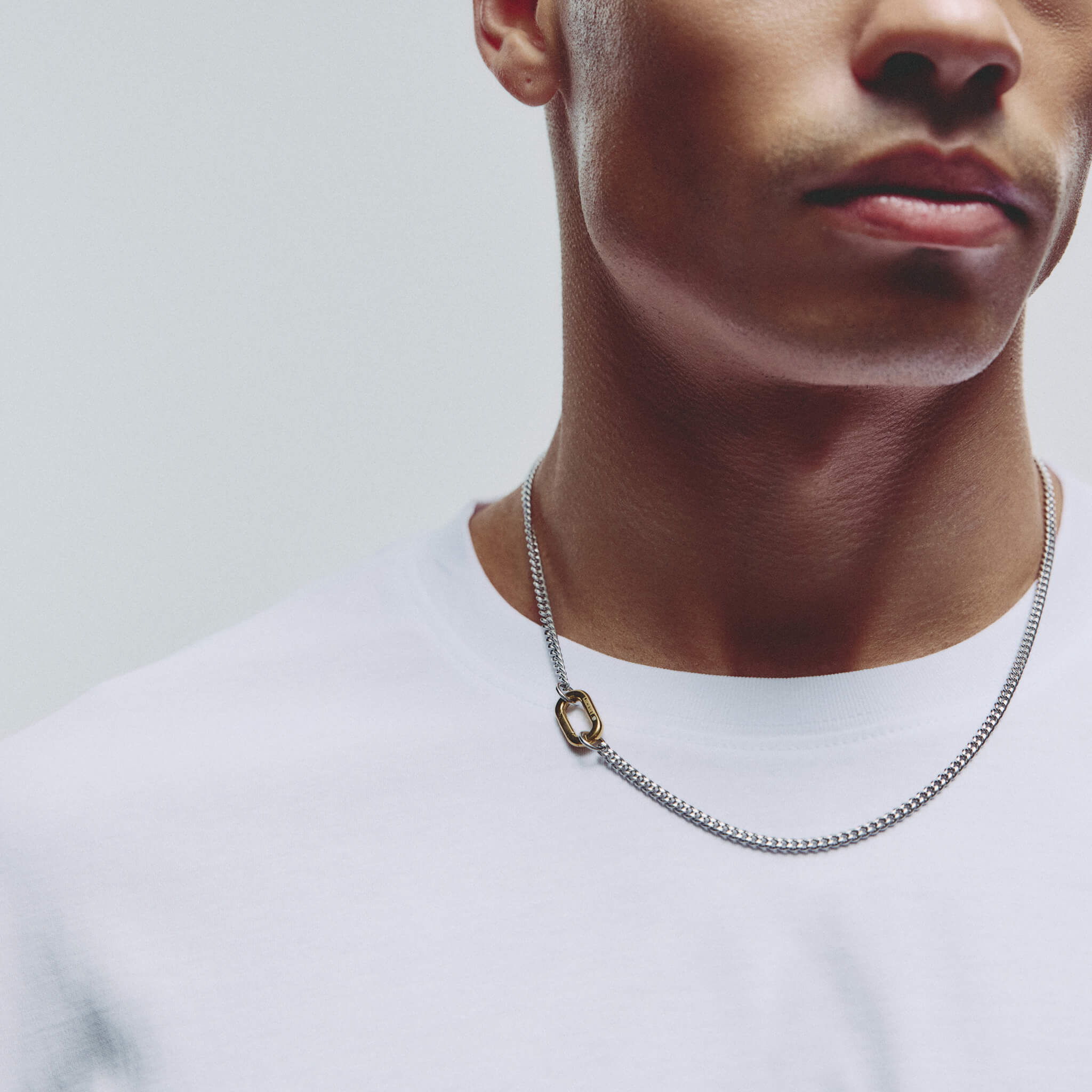 A close-up of a man wearing a silver chain necklace with a small gold carabiner clasp paired with a white crew-neck t-shirt against a light gray background.