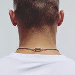 A close-up back view of a man's neck and upper shoulders, wearing a delicate silver chain necklace with a small carabiner clasp against a light gray background.