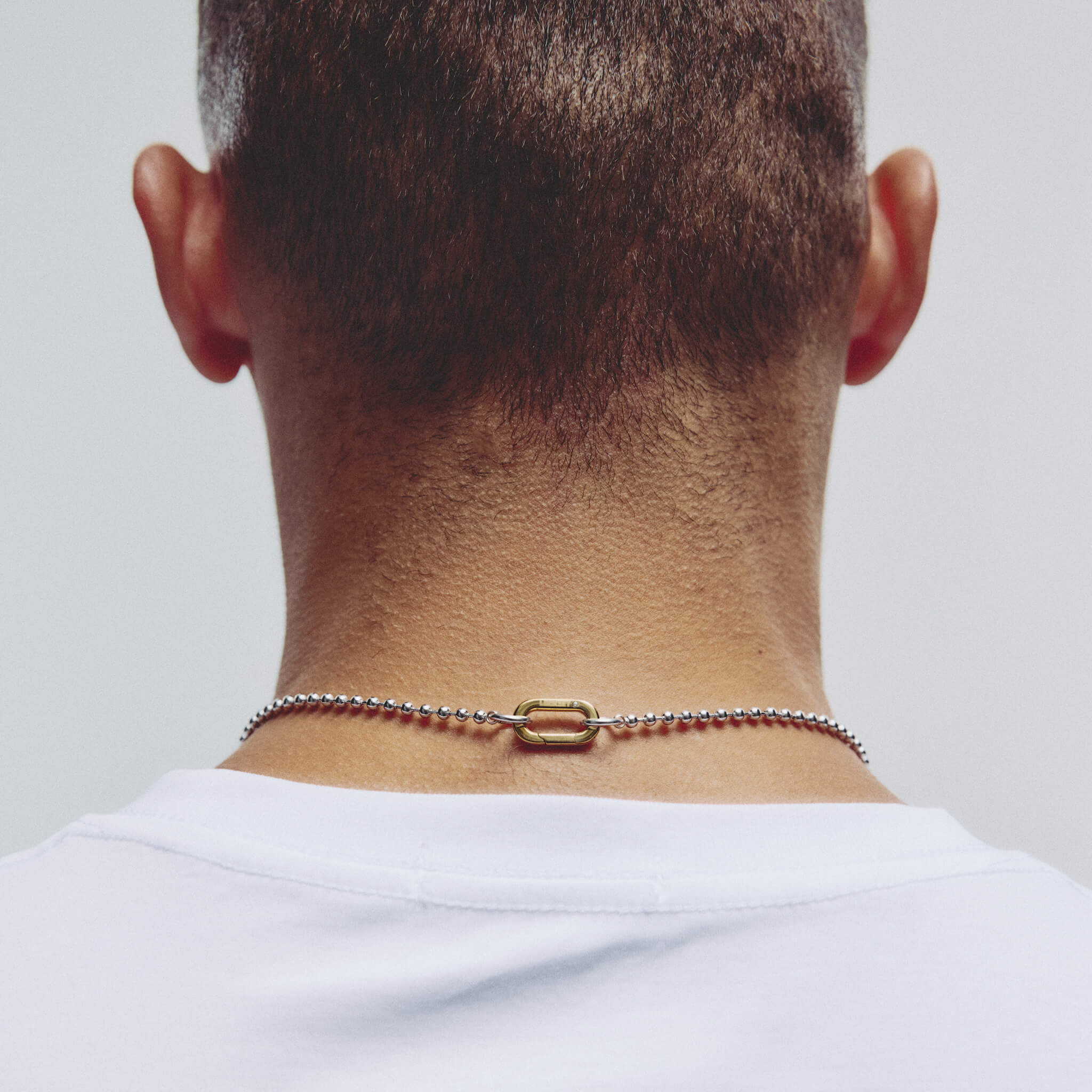 A close-up back view of a man's neck and upper shoulders, wearing a delicate silver chain necklace with a small carabiner clasp against a light gray background.