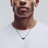 A close-up of a man wearing a silver chain necklace with 5 small geometric pendant beads, paired with a white crew-neck t-shirt against a light gray background.