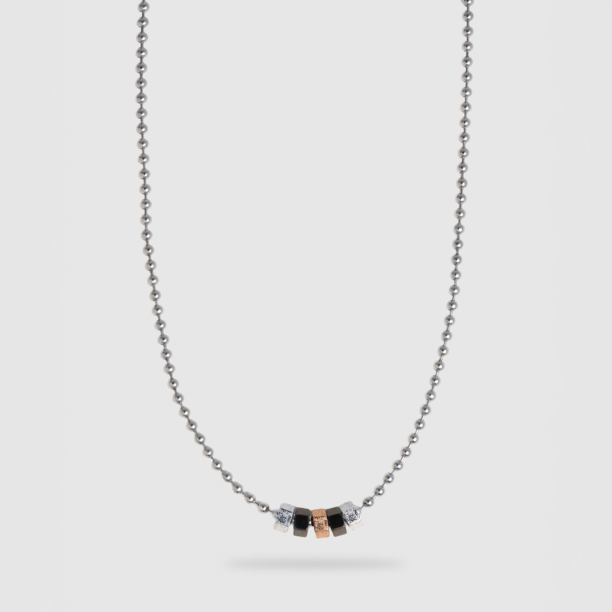 A silver ball chain necklace with a central pendant featuring five small hexagonal beads in silver, black, and rose gold, displayed against a light gray background.