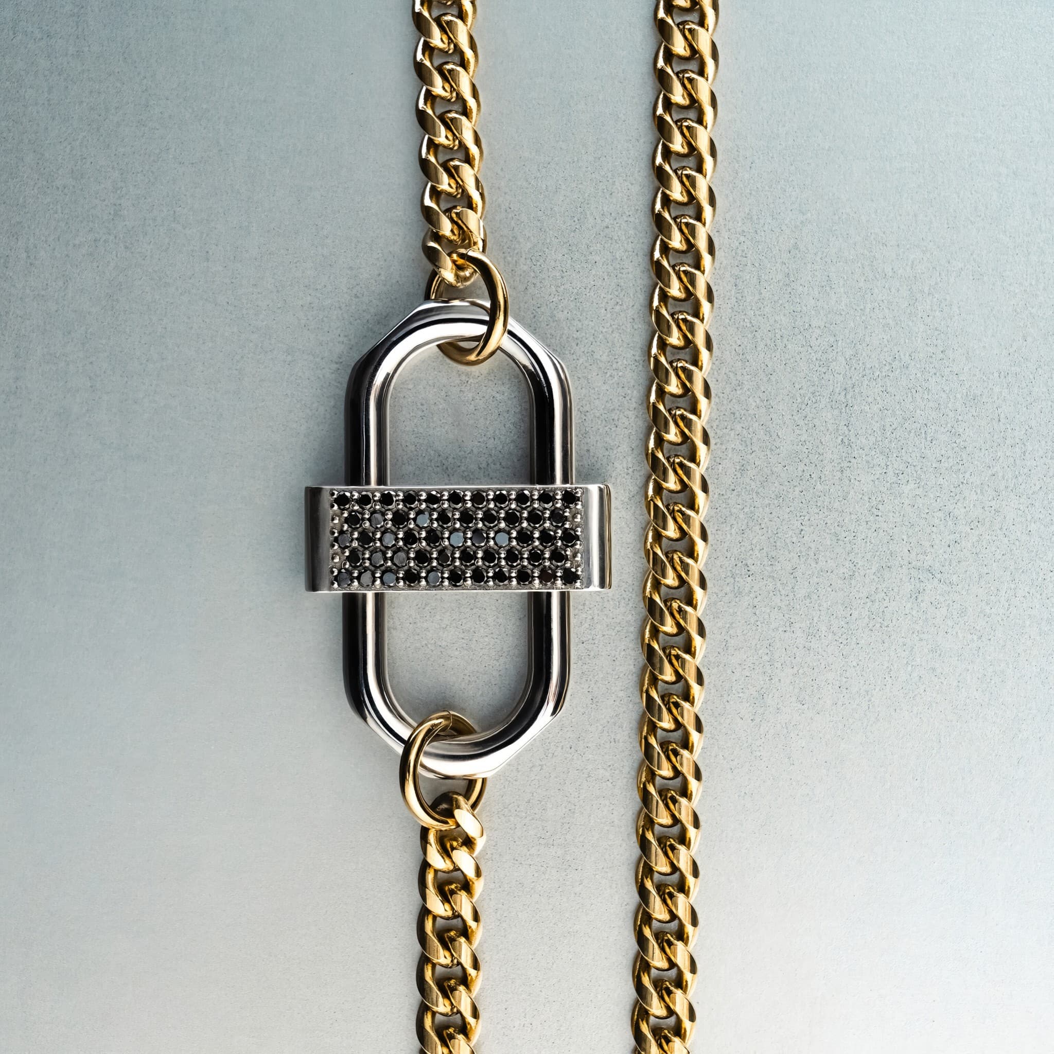 Gold chain necklace with silver double-lock pendant featuring centered black diamond detail on gray background