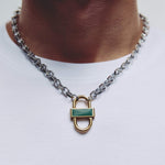 A close-up of a silver chain necklace with a padlock pendant featuring a green inlay, worn on a person's neck over a white t-shirt.