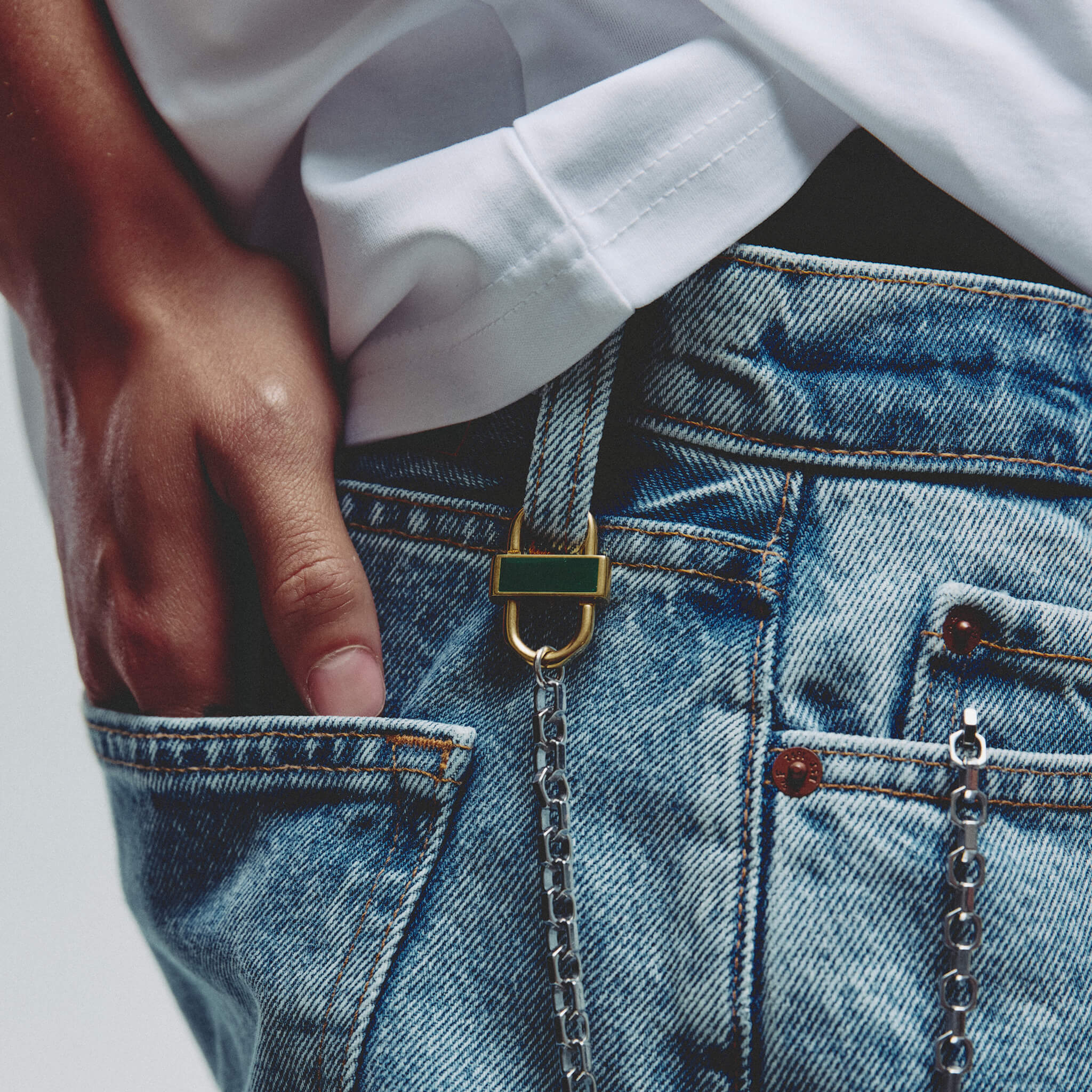 Close-up of a person wearing blue denim jeans with a gold and green enamel lock accessory attached to belt loop and silver chain.  