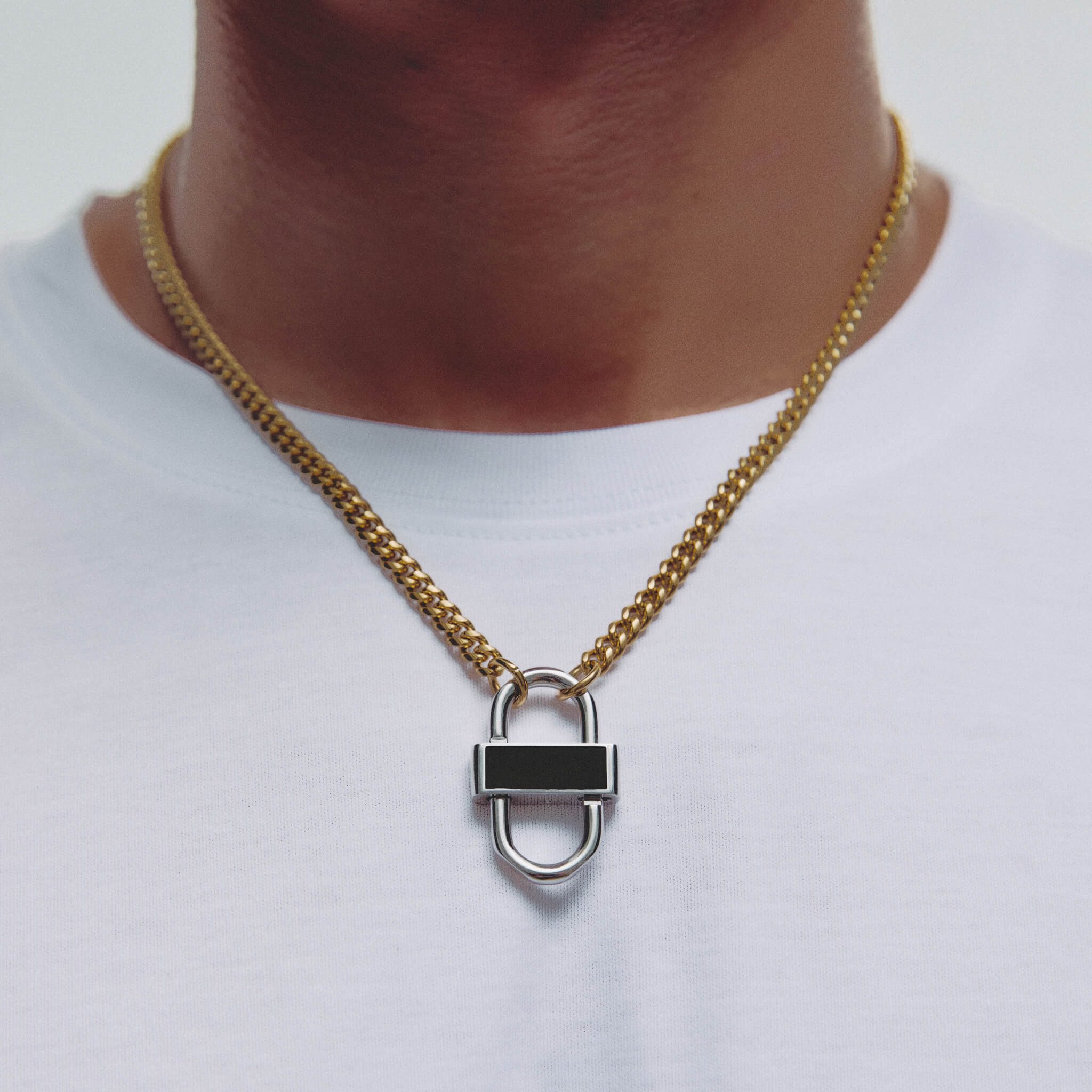 A gold chain necklace with a silver padlock pendant featuring a black enamel center, worn on a person's neck over a white t-shirt.