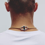 A close-up back view of a man wearing a gold chain necklace with a silver-toned lock clasp and a black rectangular inlay, paired with a white t-shirt against a light gray background.