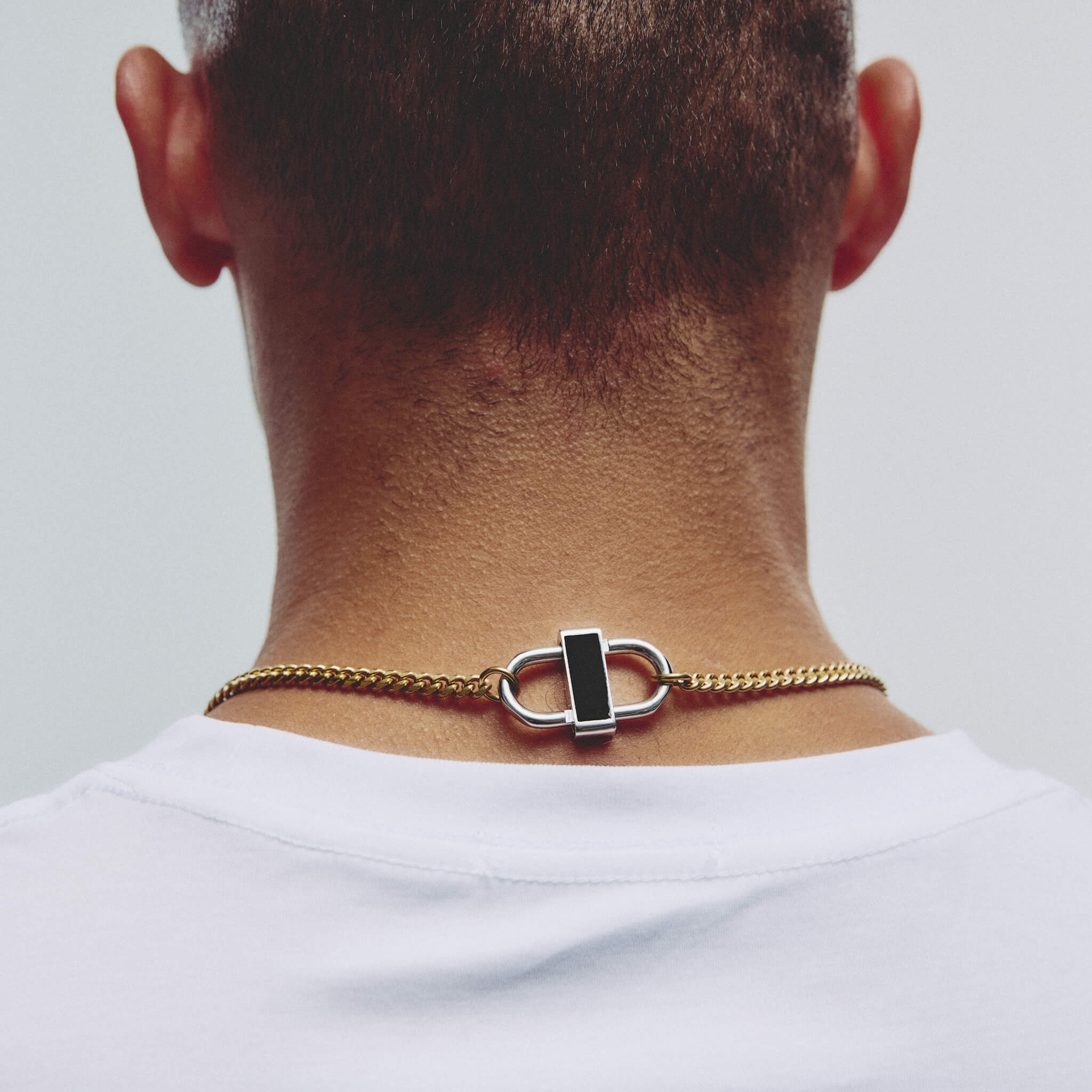 A close-up back view of a man wearing a gold chain necklace with a silver-toned lock clasp and a black rectangular inlay, paired with a white t-shirt against a light gray background.