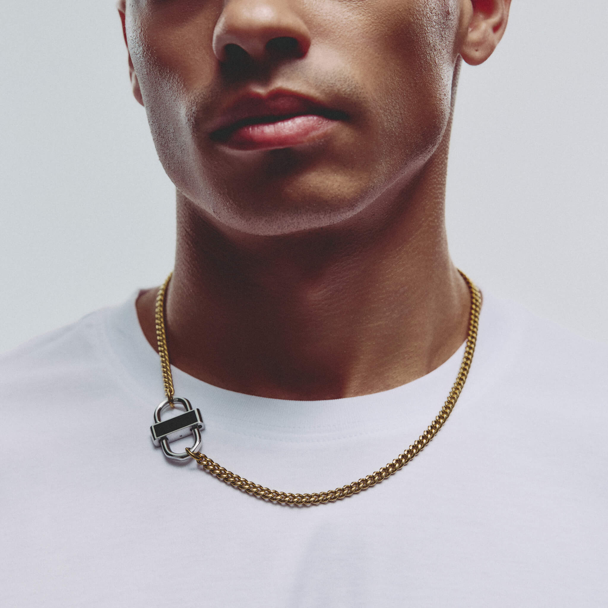 A close-up of a man wearing a gold chain necklace with a silver-toned interlocking rings pendant, styled with a white crew-neck t-shirt against a light gray background.