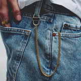 A close-up of a person wearing blue denim jeans with a gold-tone chain accessory hanging from a silver lock on a belt loop. with a hand holding the waistband.