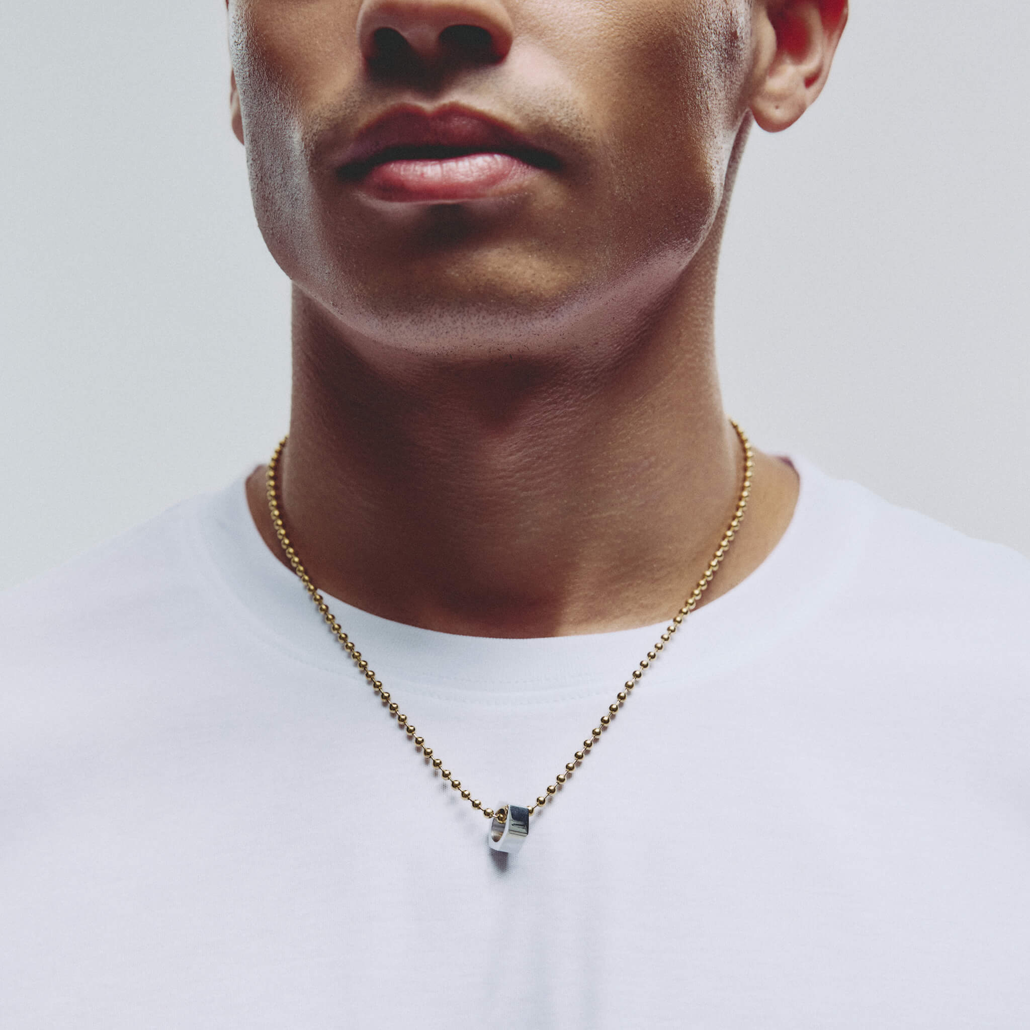 A close-up of a man wearing a gold chain necklace with a small silver hexagonal pendant, against a light gray background.