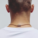 A close-up back view of a man's neck and upper shoulders, wearing a delicate gold chain necklace with a central silver carabiner clasp against a plain light gray background.