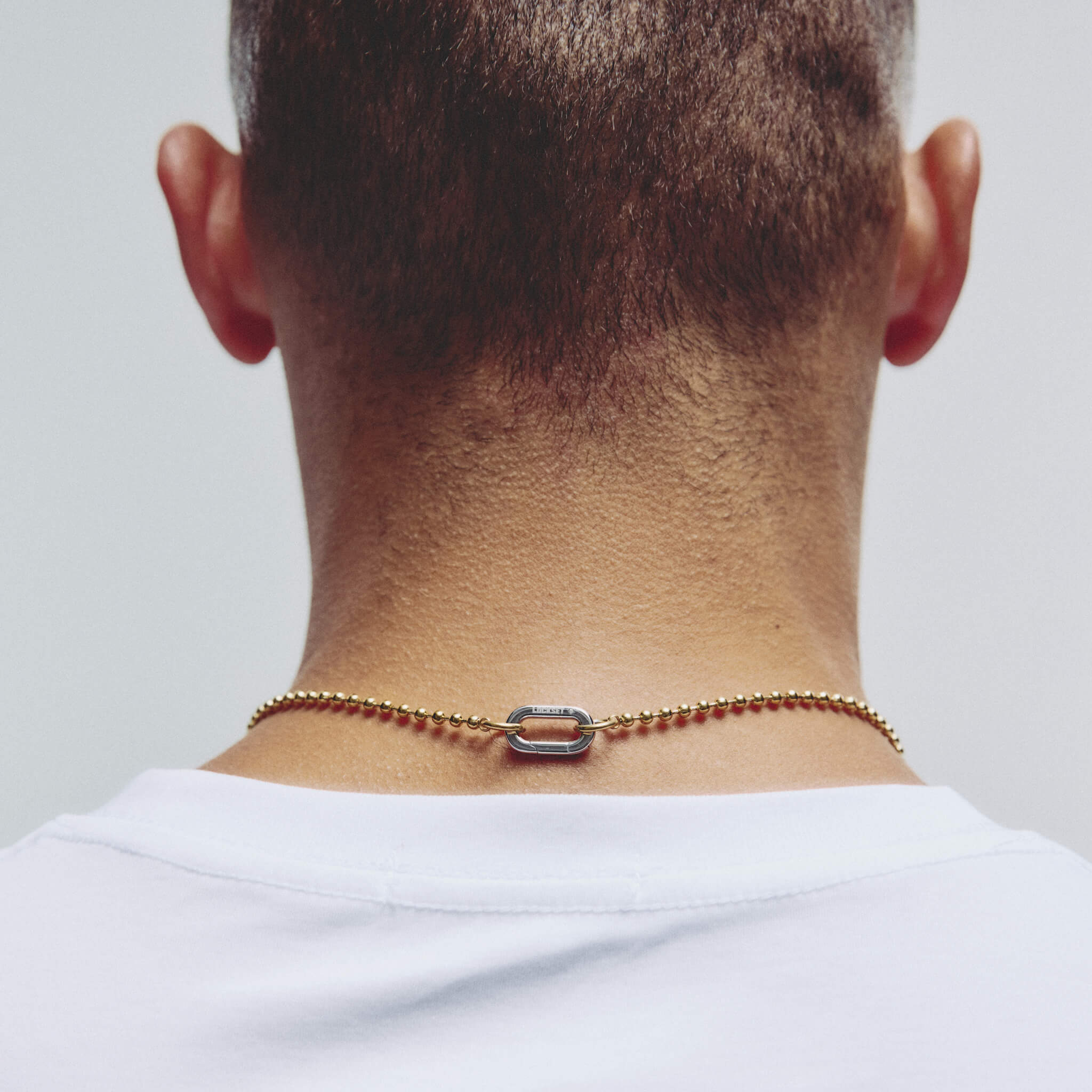 A close-up back view of a man's neck and upper shoulders, wearing a delicate gold chain necklace with a central silver carabiner clasp against a plain light gray background.