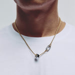 A close-up of a person's neck and upper chest wearing a delicate gold chain necklace with a small hexagonal pendant and a carabiner clasp, paired with a white t-shirt.