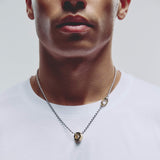 A close-up of a man wearing a silver ball chain necklace with a gold-toned hex shaped pendant, paired with a white t-shirt against a light gray background.