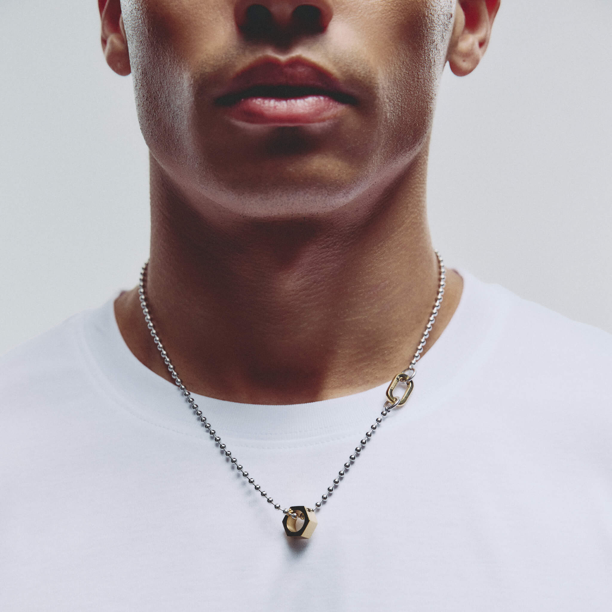 A close-up of a man wearing a silver ball chain necklace with a gold-toned hex shaped pendant, paired with a white t-shirt against a light gray background.