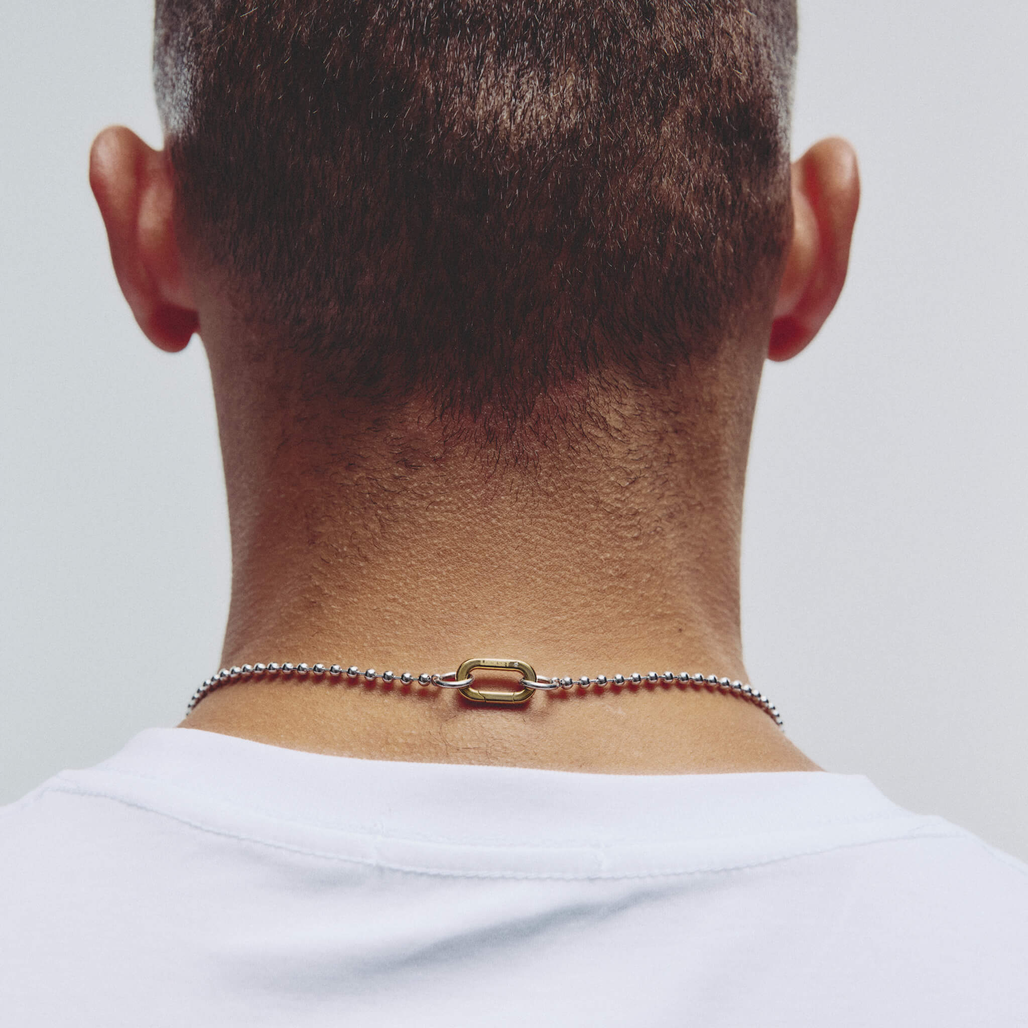 A close-up back view of a person wearing a delicate metallic chain necklace with a rectangular clasp, shown against a plain light gray background.