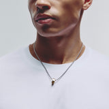 A close-up of a man wearing a silver ball chain necklace with a small gold hexagonal pendant, styled with a white crew-neck t-shirt against a light gray background.
