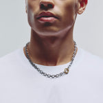 A close-up of a man wearing a silver washer shaped chain necklace, with a small gold washer shaped clasp, paired with a white t-shirt against a light gray background.
