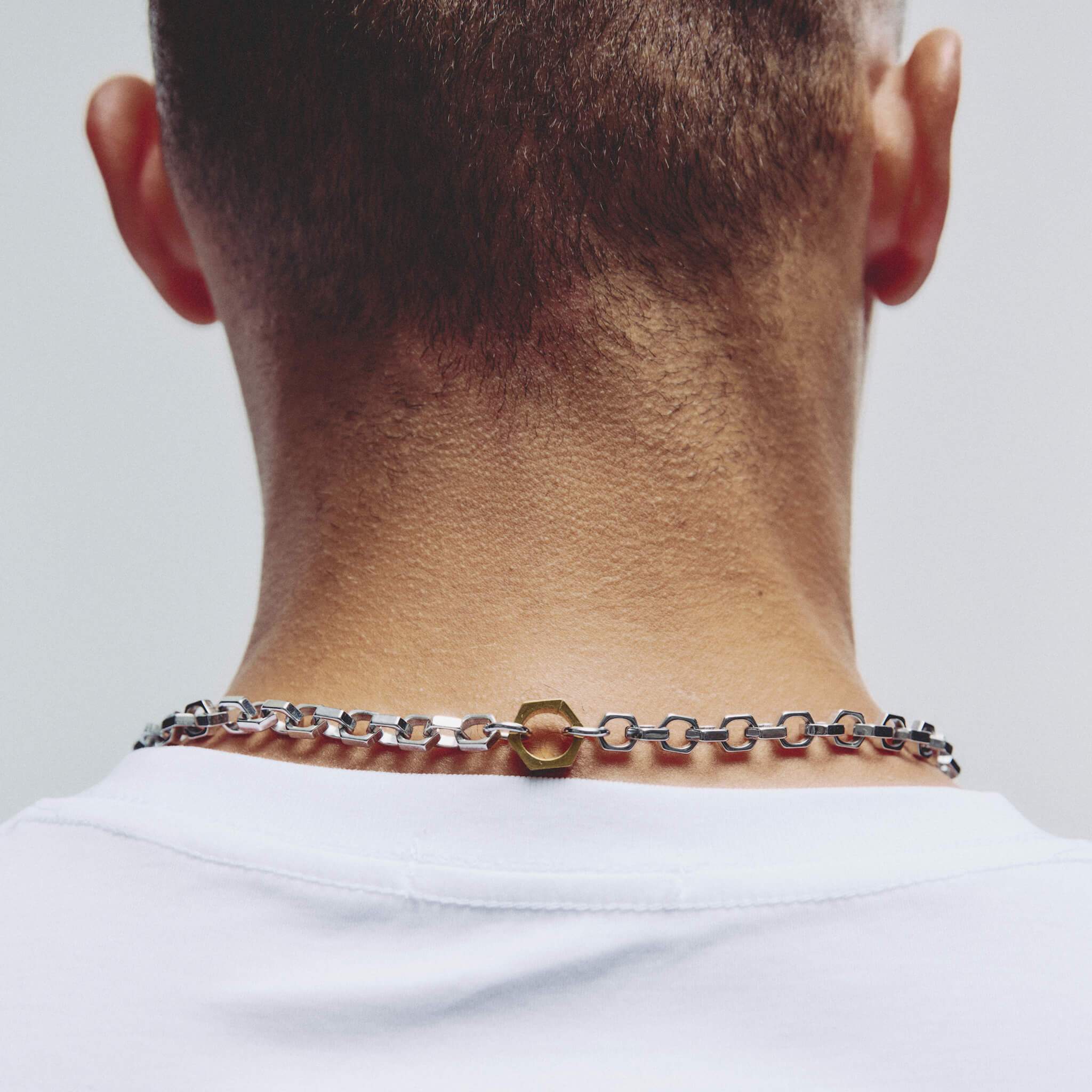 A close-up back view of a man's neck and upper shoulders wearing a silver-toned chain necklace with a central gold washer clasp, over a white t-shirt.