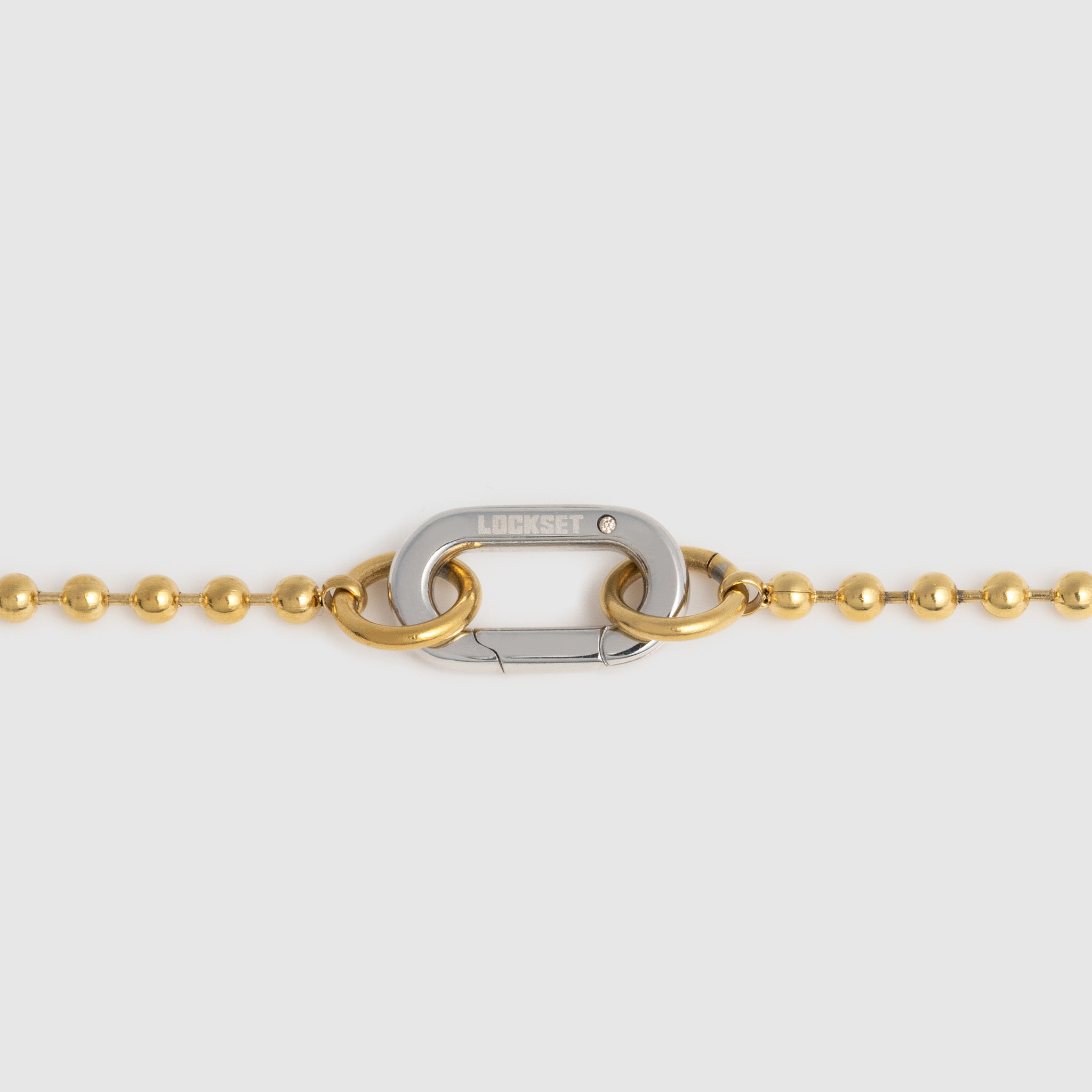A close-up of a gold ball chain necklace featuring a central silver carabiner clasp with the brand name 'LOCKSET' engraved on it, set against a light gray background.