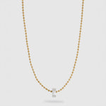 A delicate gold ball chain necklace with a small, polished silver hexagonal pendant, displayed against a light gray background.