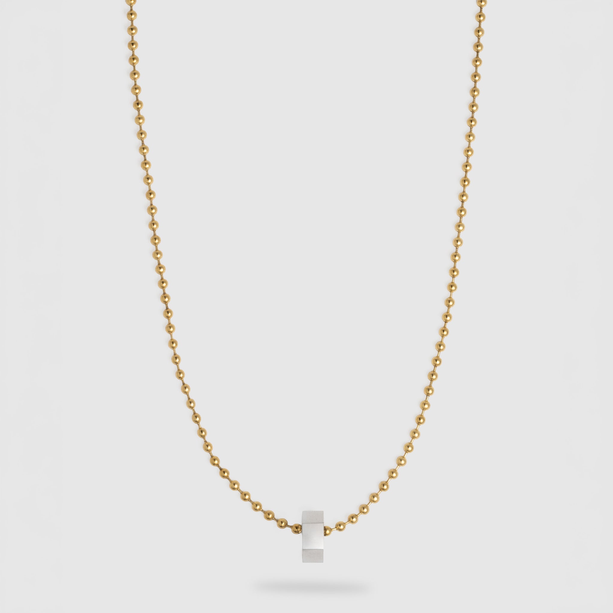 A delicate gold ball chain necklace with a small, polished silver hexagonal pendant, displayed against a light gray background.