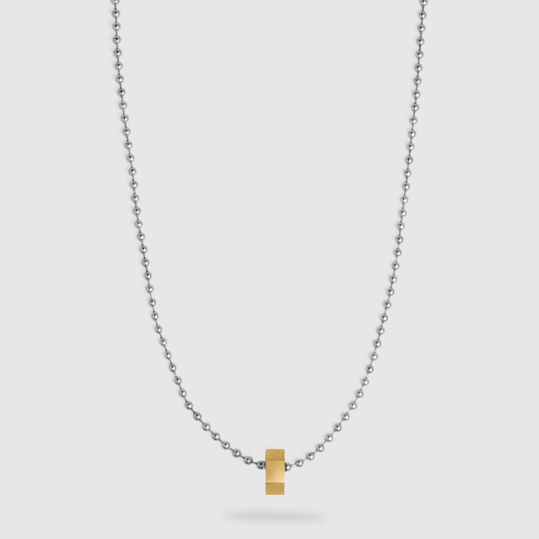 A delicate silver ball chain necklace featuring a small, polished gold hexagonal pendant at the center, displayed against a light gray background.