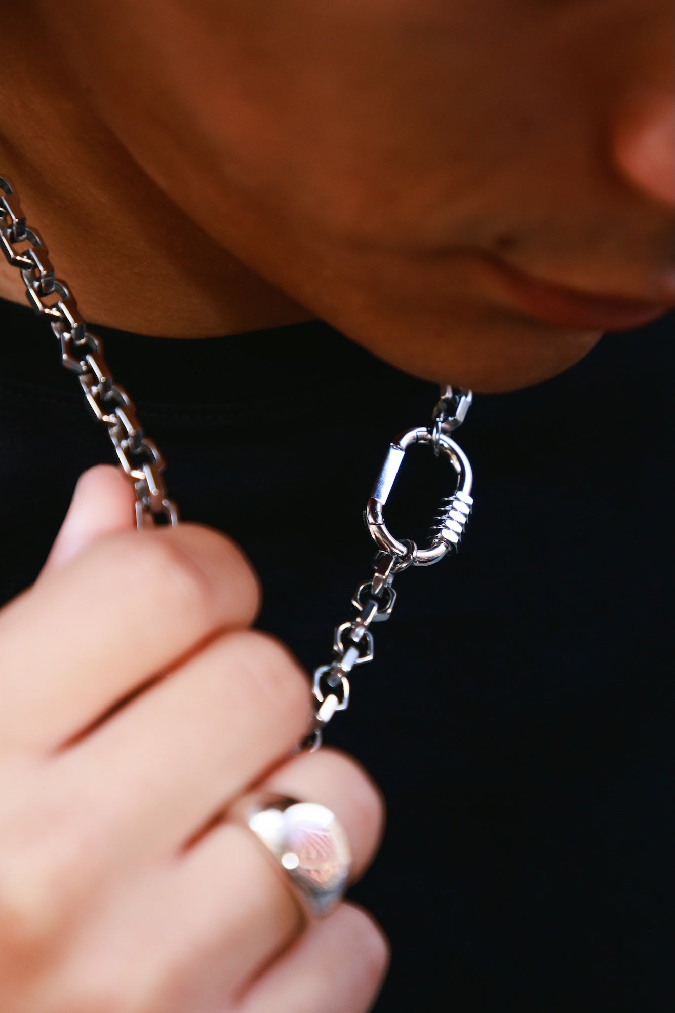 A close-up lifestyle shot of a person wearing a silver chain necklace with a carabiner clasp, holding the chain near their neck against a black background.