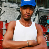 A man wearing a blue Lockset baseball cap and a white tank top stands with arms crossed in front of a graffiti-covered wall.