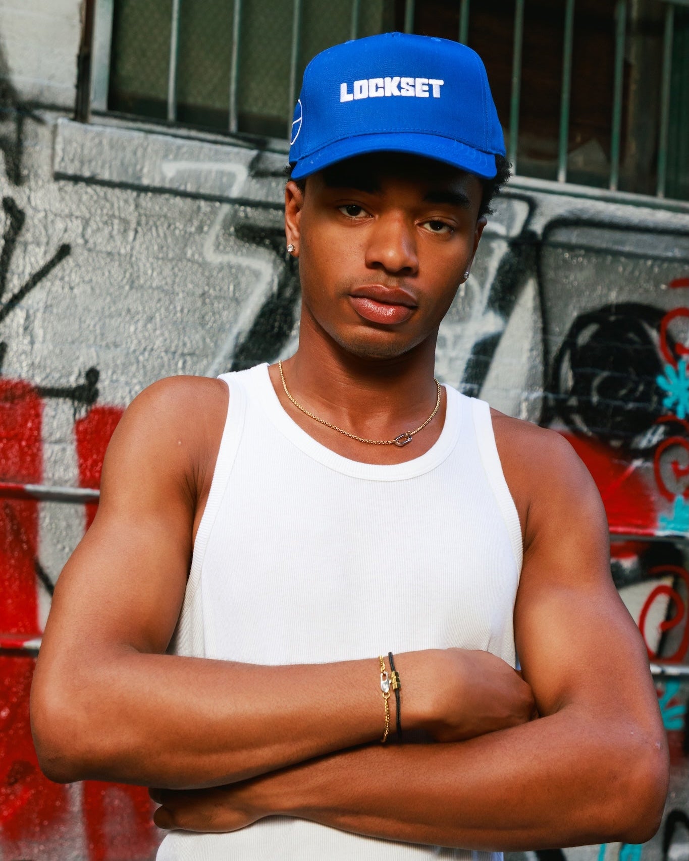 A man wearing a blue Lockset baseball cap and a white tank top stands with arms crossed in front of a graffiti-covered wall.