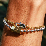 A close-up of a person's wrist wearing two bracelets: one gold chain bracelet with a hex shaped clasp and one silver cuban bracelet with a clasp.