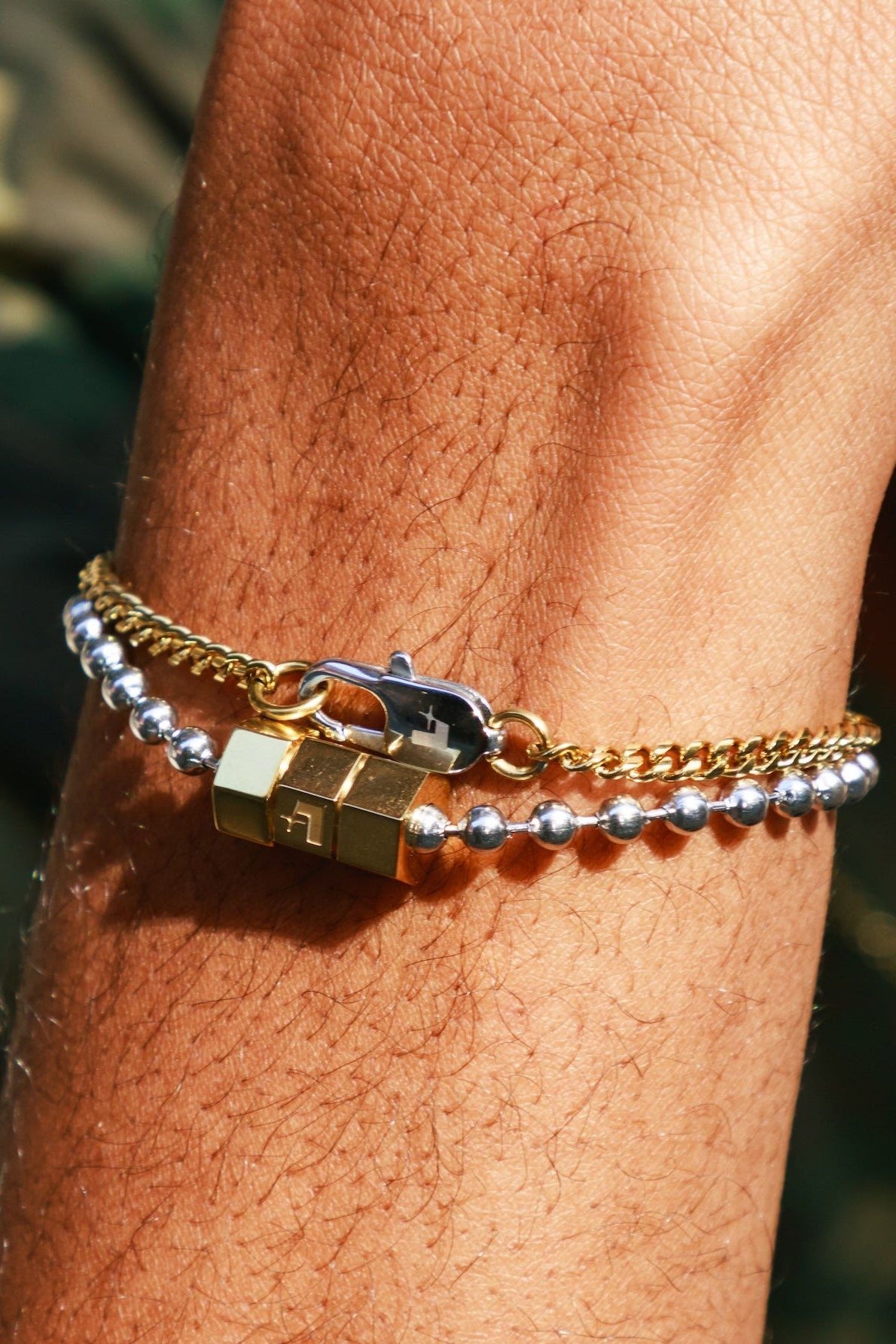 A close-up of a person's wrist wearing two bracelets: one gold chain bracelet with a hex shaped clasp and one silver cuban bracelet with a clasp.