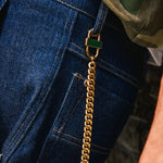 Close-up of a gold Cuban chain styled as a pant chain, attached to a gold lock and looped through the belt loop of dark blue denim jeans, with a camouflage shirt partially visible.