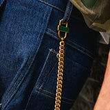 Close-up of a gold Cuban chain styled as a pant chain, attached to a gold lock and looped through the belt loop of dark blue denim jeans, with a camouflage shirt partially visible.