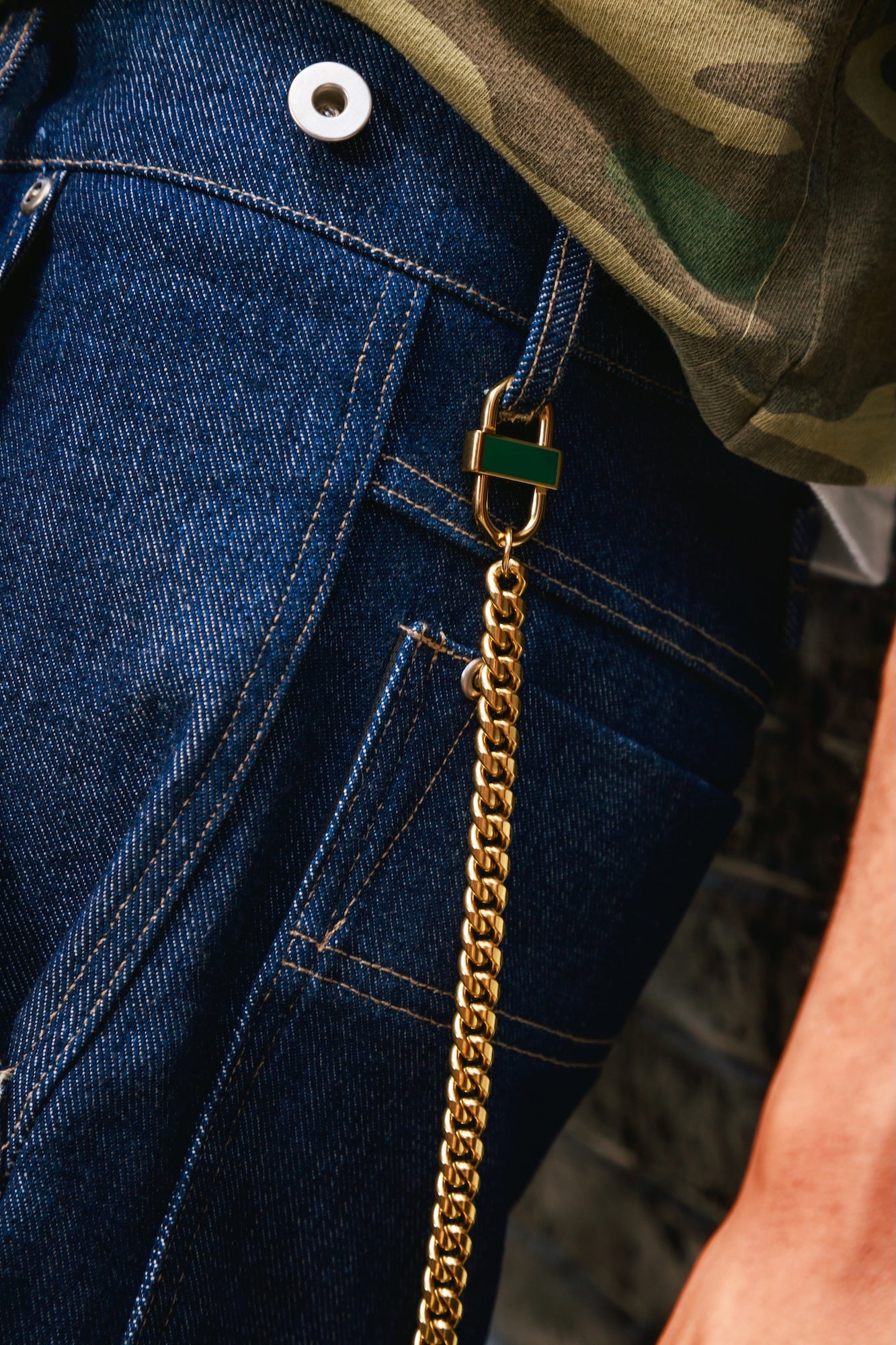 Close-up of a gold Cuban chain styled as a pant chain, attached to a gold lock and looped through the belt loop of dark blue denim jeans, with a camouflage shirt partially visible.
