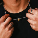 A close-up of a person wearing a silver ball-chain necklace with a small gold hex pendant, adjusting it with both hands while wearing a black t-shirt and silver rings.