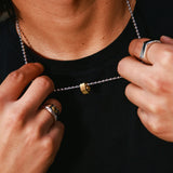 A close-up of a person wearing a silver ball-chain necklace with a small gold hex pendant, adjusting it with both hands while wearing a black t-shirt and silver rings.