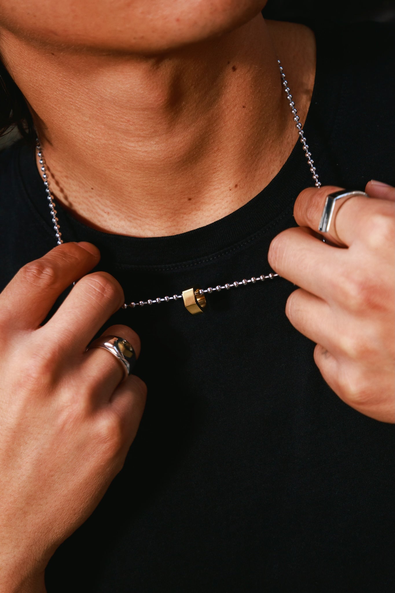 A close-up of a person wearing a silver ball-chain necklace with a small gold hex pendant, adjusting it with both hands while wearing a black t-shirt and silver rings.