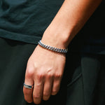 A hand against an all-black outfit, wearing a silver Cuban chain bracelet paired with a silver flat-top signet ring.