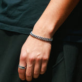 A hand against an all-black outfit, wearing a silver Cuban chain bracelet paired with a silver flat-top signet ring.
