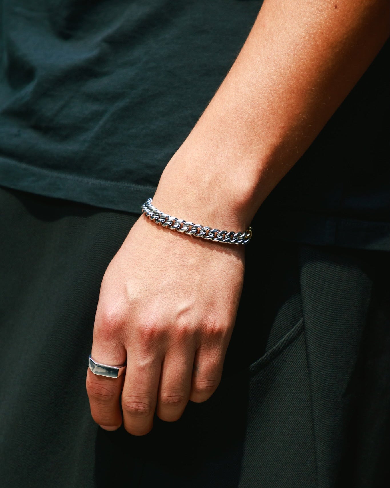 A hand against an all-black outfit, wearing a silver Cuban chain bracelet paired with a silver flat-top signet ring.