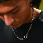 A close-up lifestyle shot of a man wearing a silver chain necklace with a small gold hexagonal pendant, looking down in warm natural light.